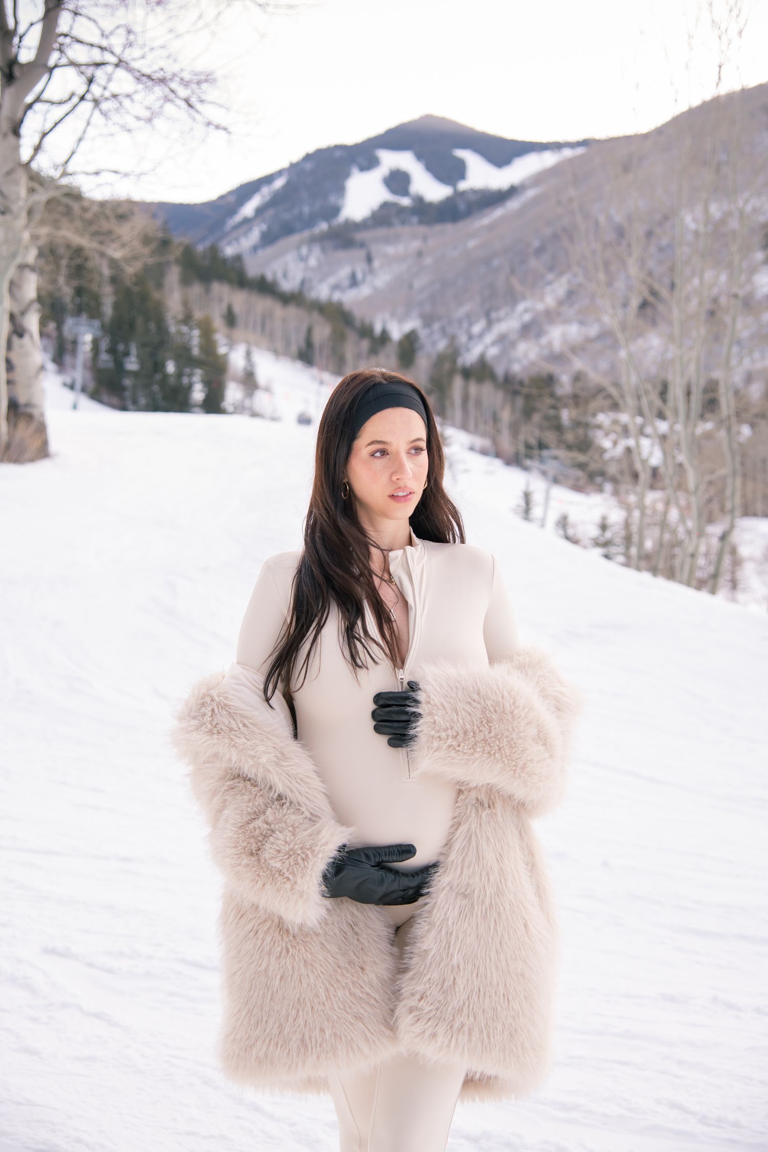 Woman in luxurious cream fur coat poses against snowy mountain landscape at winter ski resort for her fashion maternity photos in Vail CO