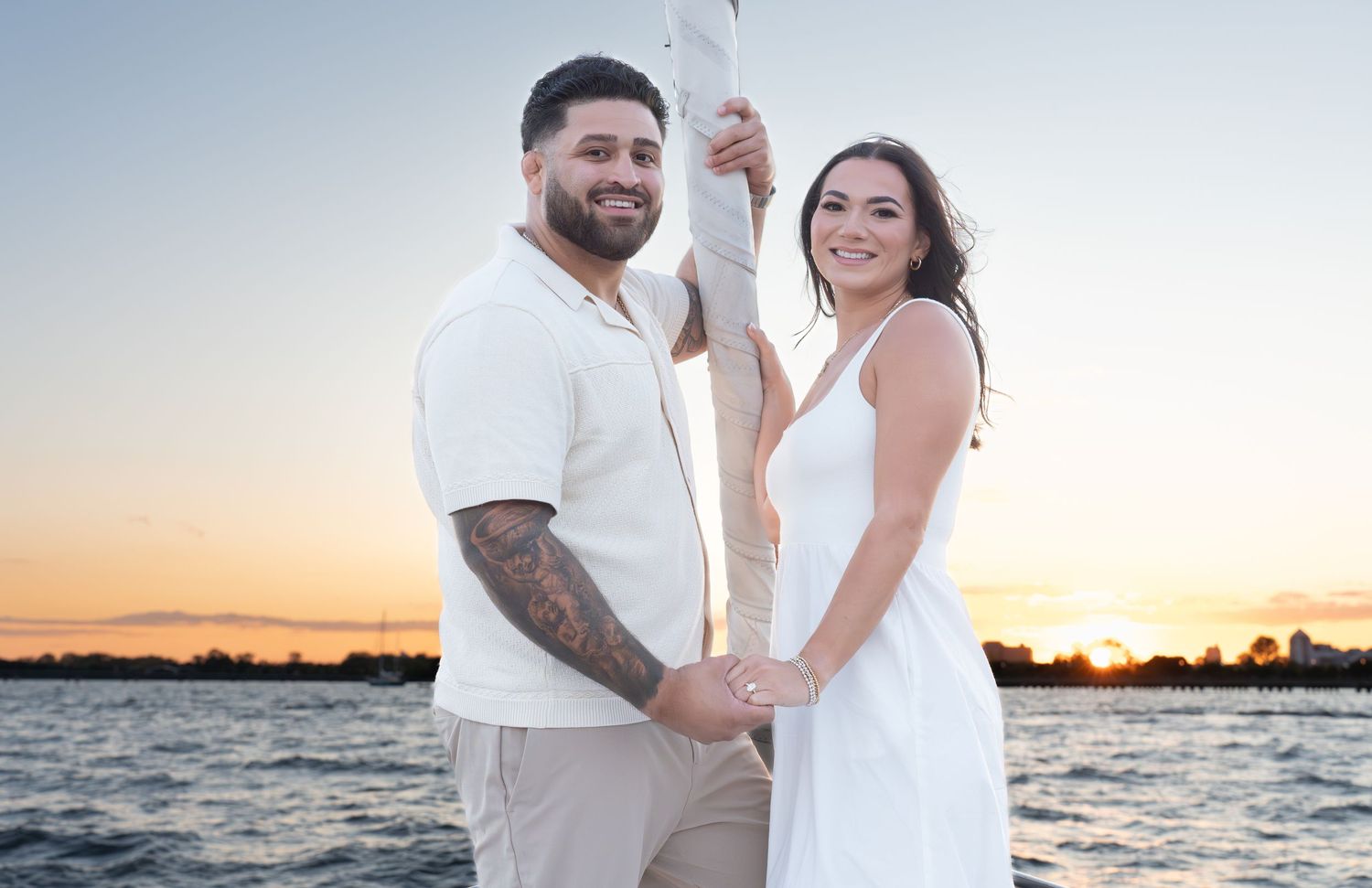 A couple in white outfits holds hands on a sailboat during sunset over calm waters.
