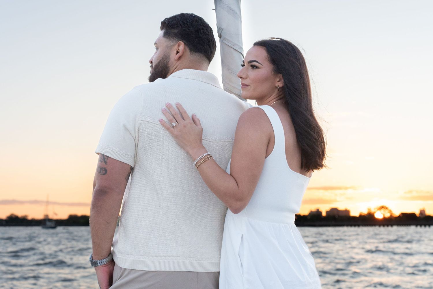 Couple in white outfits embracing by the water during sunset with orange sky in background.