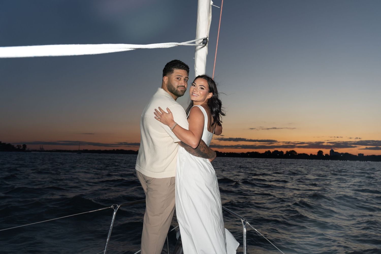 Couple embraces during a romantic sunset photoshoot by the ocean with boats in the background.