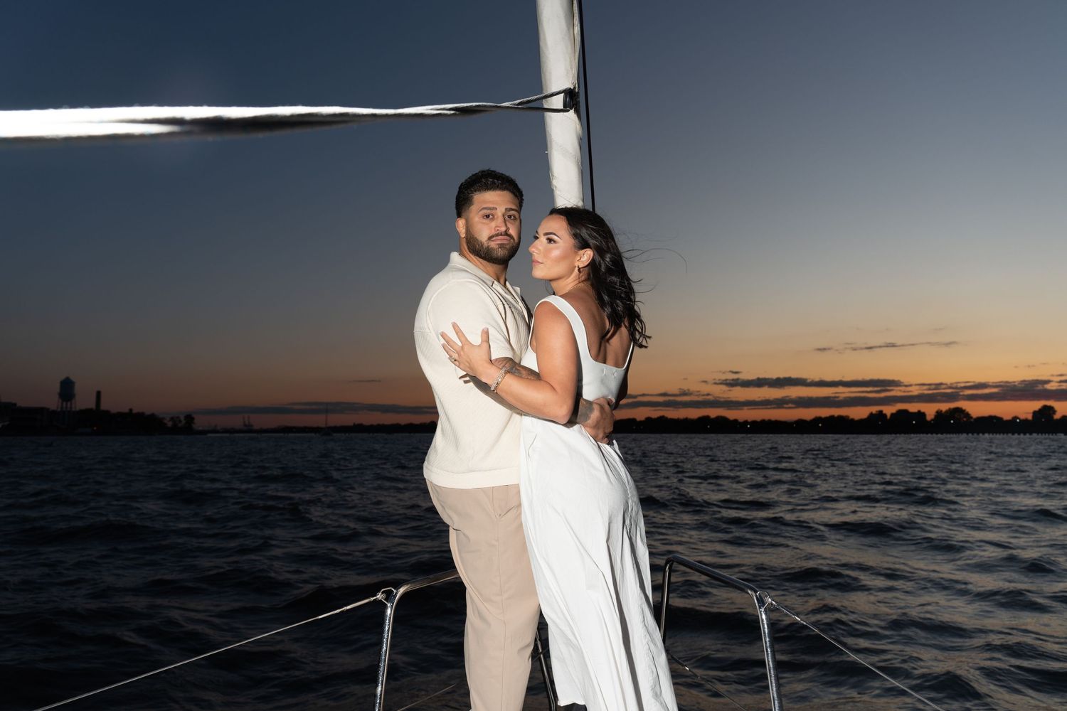 A couple embraces on a sailboat at sunset with the water and skyline in the background.