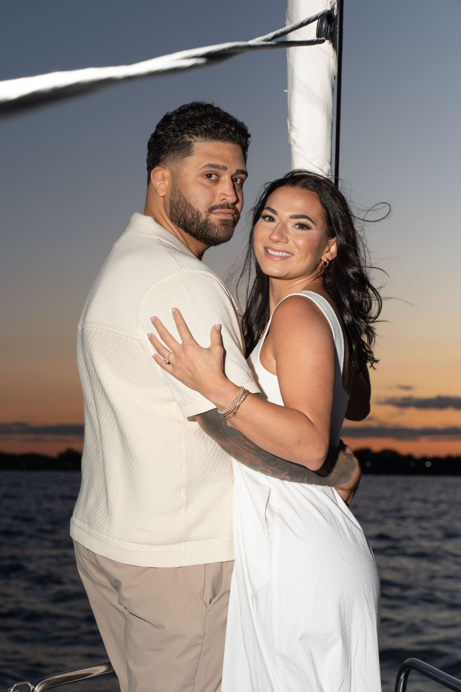 A couple embraces during sunset by the ocean in coordinated white and cream outfits for romantic photos.