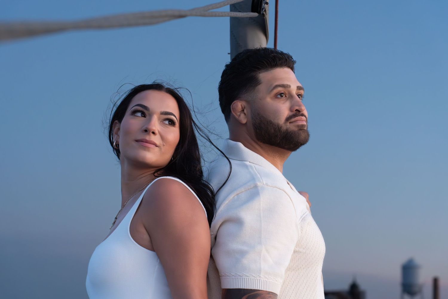 A couple in white clothing embraces against a blue sky backdrop during a romantic outdoor photoshoot.