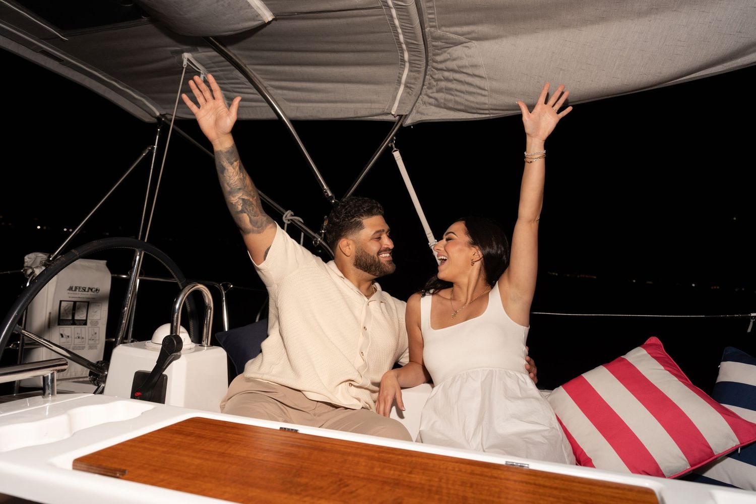 A group of people celebrate with raised arms while riding in a boat on the water at night.