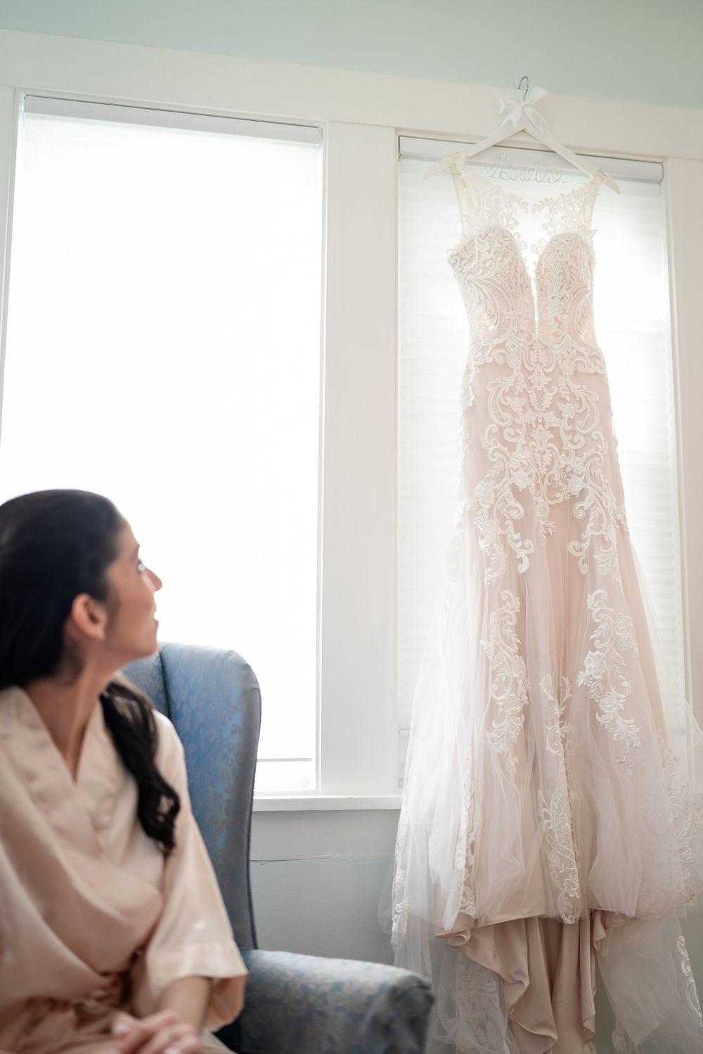 A blush pink lace wedding gown hangs elegantly by a window while someone in a robe sits nearby on a blue chair.