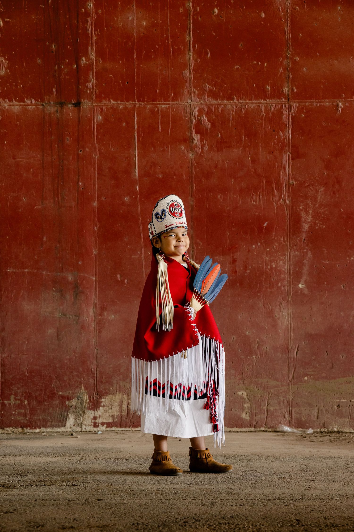 A child wearing traditional Native American regalia with fringe and beadwork stands against a rustic red wall.