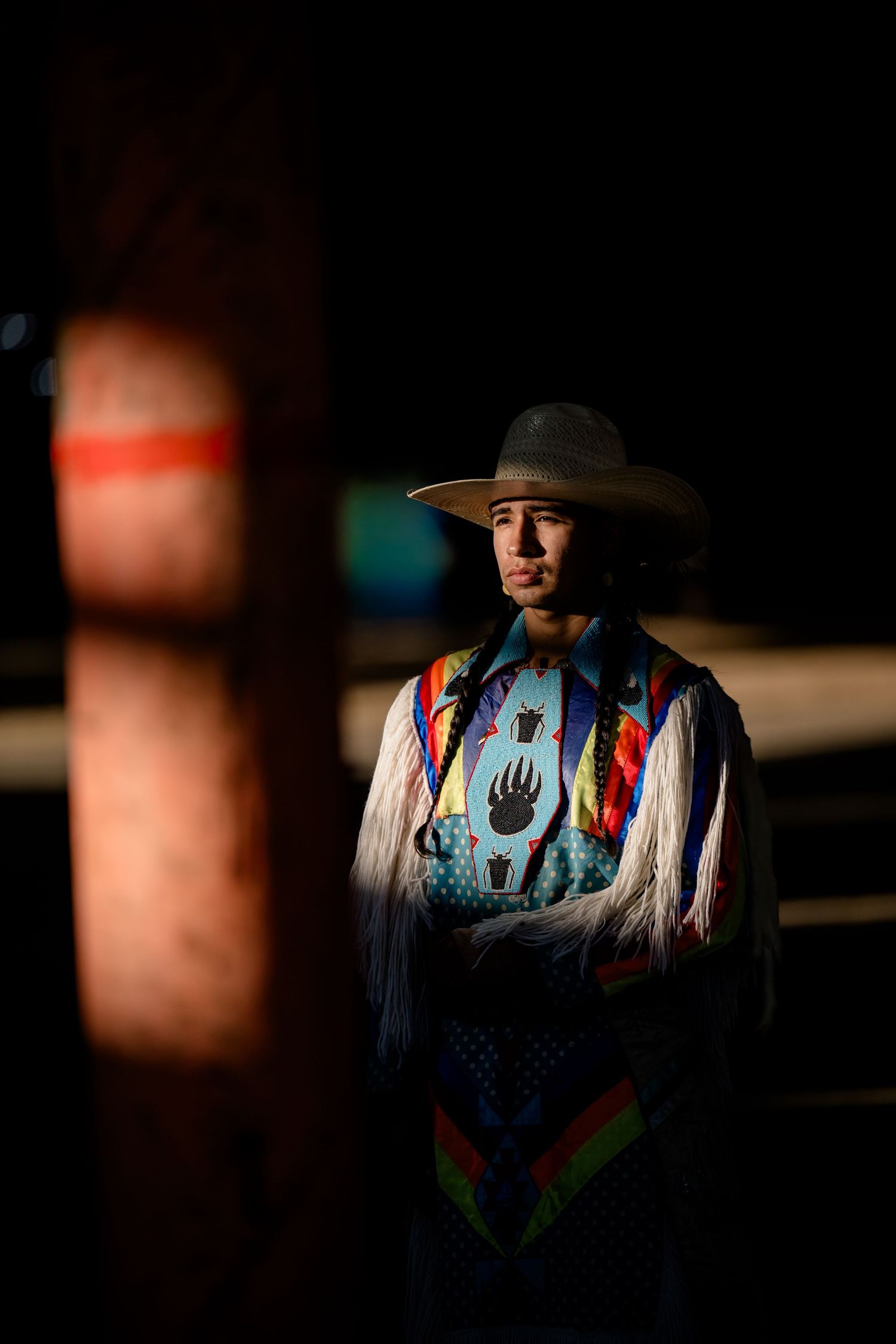 Person wearing traditional Native American regalia with a wide-brimmed hat stands in dramatic lighting.