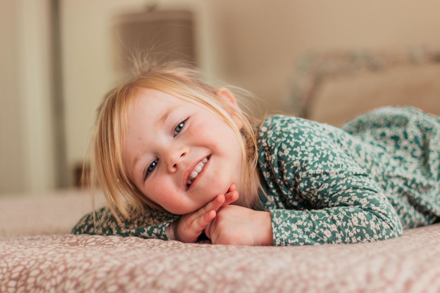 Smiling toddler girl with blonde hair lying on a bed, resting her chin on her hands in a floral top | Kids Birthday Photo Sessions