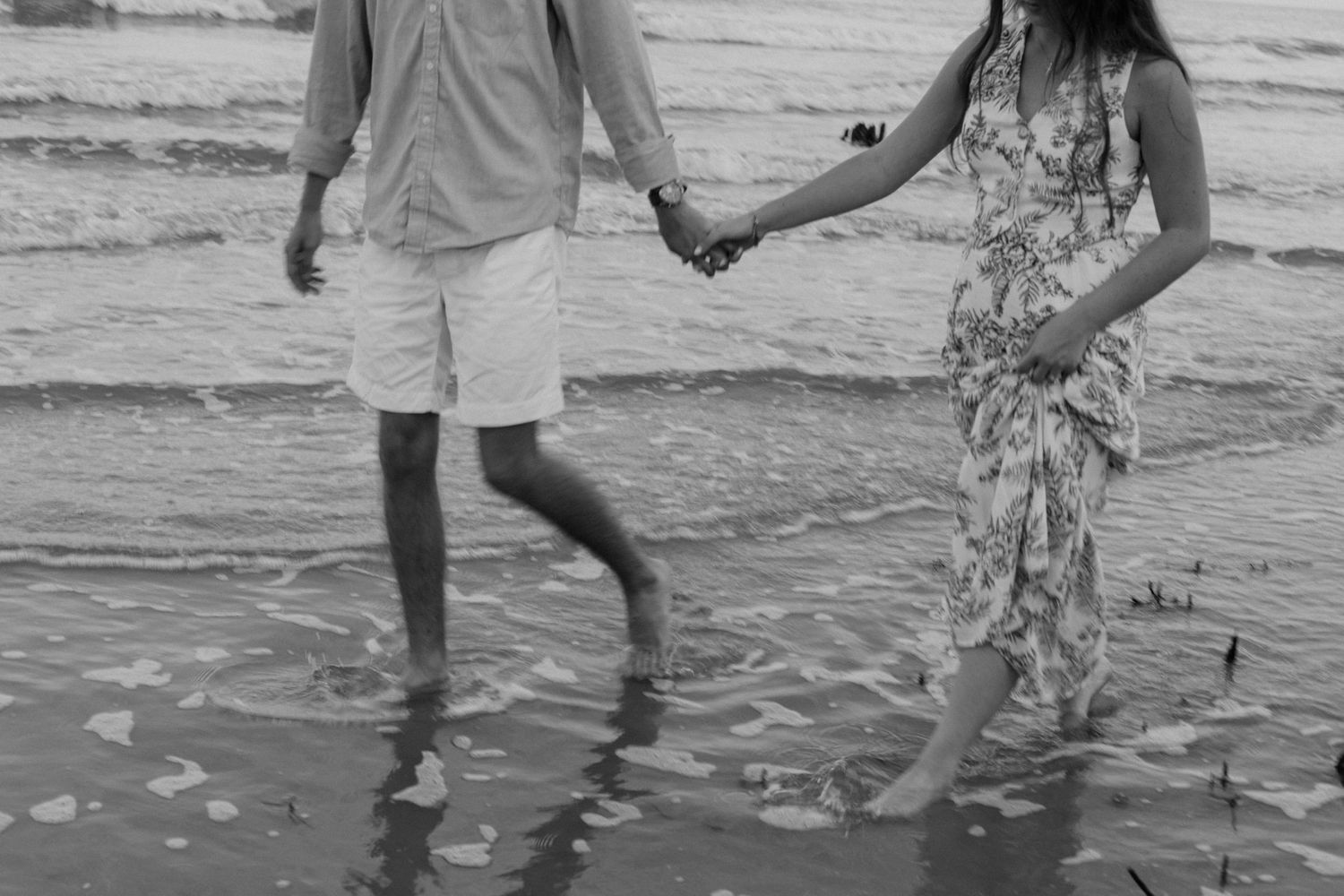 Black and white photo of couple holding hands while walking in beach surf.