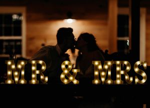 Couple silhouetted kissing behind illuminated marquee Mr and Mrs letters at wedding reception.