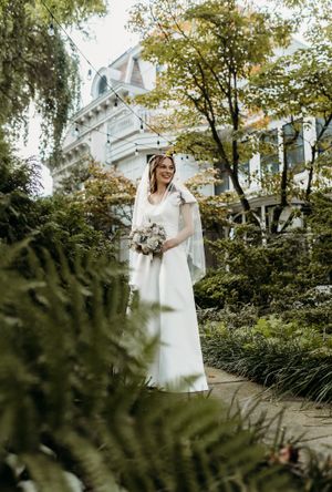 Bride in simple white gown and veil stands in lush garden path before Victorian mansion.