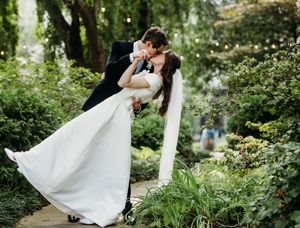 Groom dipping bride for a kiss in a lush garden with string lights overhead.