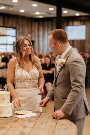Bride in lace gown and groom in gray suit share a smile while cutting their white wedding cake at reception.