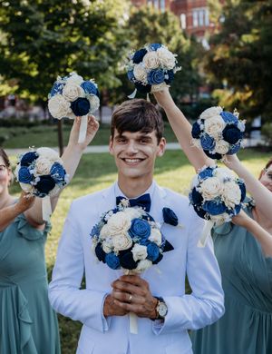 Smiling groom in white suit surrounded by bridesmaids holding blue and white bouquets.