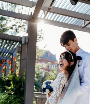 Laughing bride and groom embrace under pergola with sunlight streaming through, holding blue bouquet.