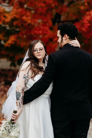 Tattooed bride in white gown with veil gazes at groom against brilliant red autumn tree backdrop.
