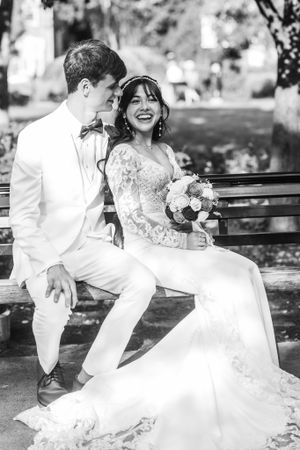 Black and white photo of a laughing bride holding bouquet sitting with groom on park bench in wedding attire.