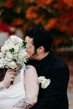 Bride and groom sharing romantic kiss hidden behind white rose and baby's breath wedding bouquet.