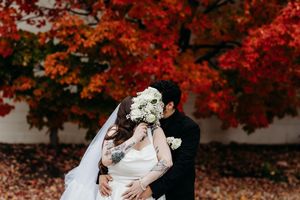 Bride and groom sharing a kiss behind white bouquet with vibrant red autumn foliage backdrop.