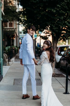 Bride and groom in white wedding attire holding hands on urban sidewalk smiling back at camera.