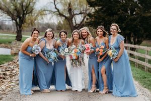 Bride poses with six bridesmaids in dusty blue gowns holding colorful bouquets on a gravel path at spring wedding.