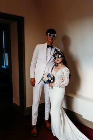 Bride and groom posing in white outfits wearing heart-shaped sunglasses indoors.