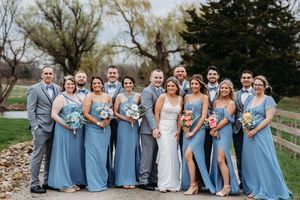 Wedding party poses together, bridesmaids in blue gowns and groomsmen in gray suits, holding colorful bouquets outdoors.