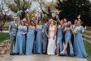 Joyful wedding party celebrates with raised bouquets, bridesmaids in blue dresses and groomsmen in gray suits kissing couple.