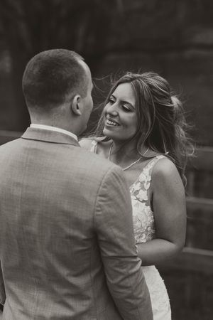 Black and white portrait of smiling bride gazing at groom in gray suit during outdoor wedding ceremony.