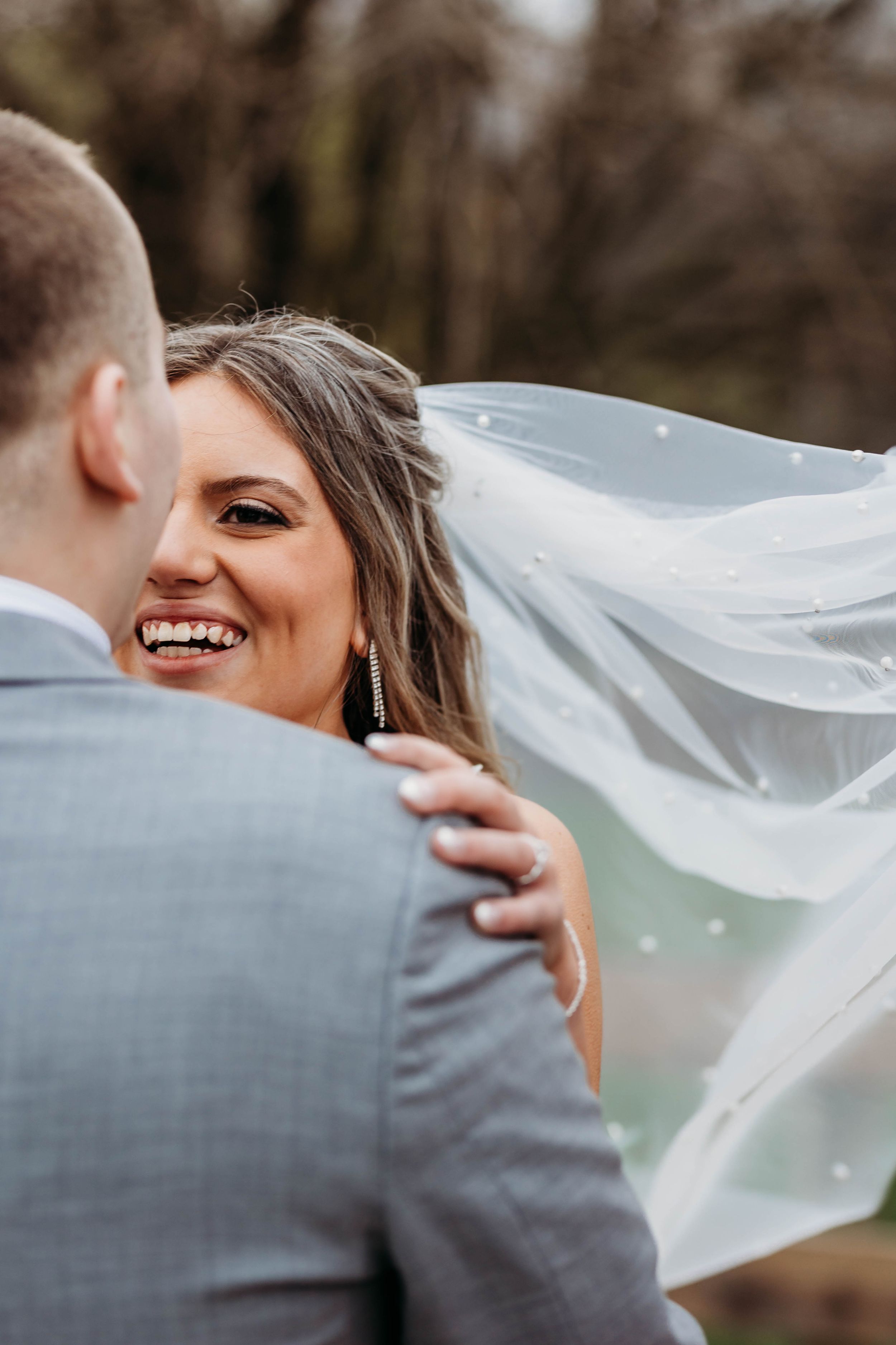 Laughing bride with flowing veil blown by wind embraces groom in gray suit during outdoor wedding portraits.