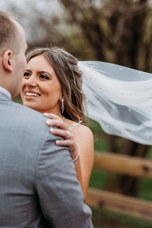Radiant bride smiling at groom while her pearl-adorned veil flows in the wind during wedding portraits.