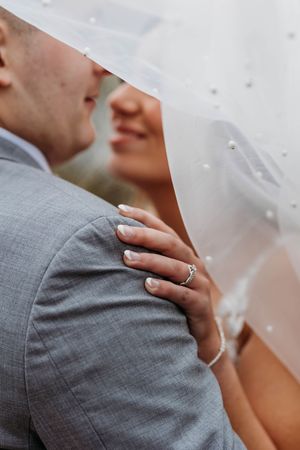 Close-up of bride resting hand on grooms shoulder showing diamond ring under flowing pearl wedding veil.