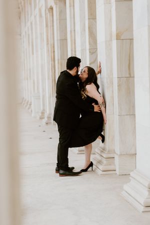 Couple sharing passionate kiss leaning against marble columns in elegant colonnade.