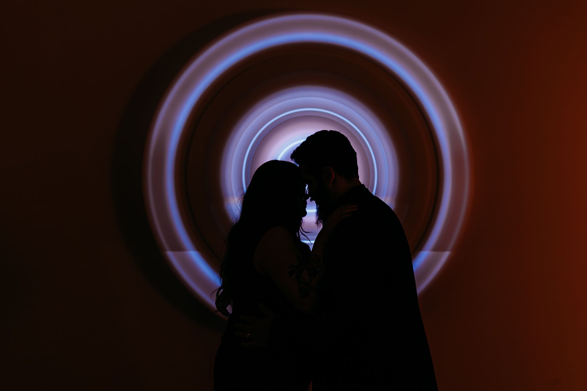 Romantic couple silhouette kissing against glowing circular light installation backdrop.