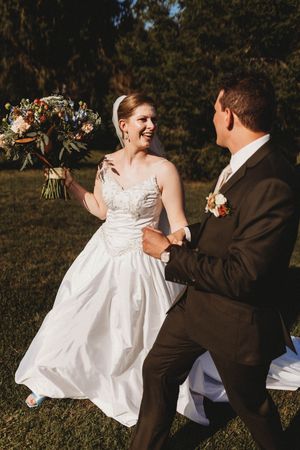 Joyful bride in lace gown holding bouquet laughs while dancing with groom in black suit outdoors.