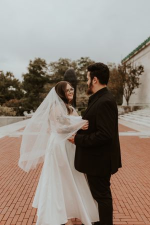 Bride in white gown and veil exchanges vows with groom in black suit outdoors on brick plaza.