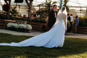 Bride with long cathedral veil stands with groom near greenhouse garden at dusk wedding reception.