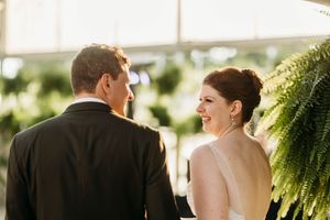 Bride smiling at groom during wedding ceremony surrounded by lush green ferns in a greenhouse venue.