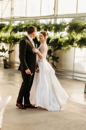 Bride and groom share a romantic moment dancing in a sunlit greenhouse with hanging plants.
