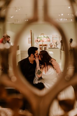 Bride and groom share a kiss reflected in an ornate mirror inside an elegant museum venue.