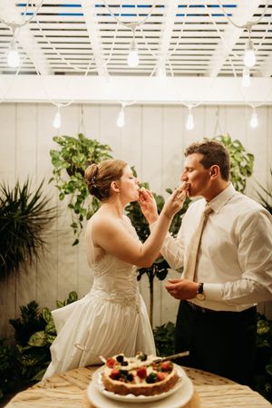 Bride and groom feed each other wedding cake decorated with fresh berries under string lights.