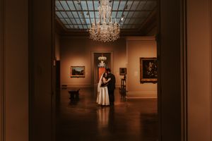 Couple embracing in a dimly lit art museum gallery beneath a crystal chandelier.