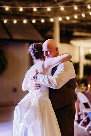 Bride shares an emotional father-daughter dance embrace under warm string lights at wedding reception.