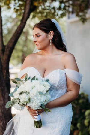 Elegant bride in off-shoulder lace gown holding white hydrangea bouquet standing under tree outdoors.