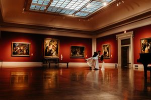 Bride in flowing white dress twirls in a grand museum gallery with red walls and classic paintings.