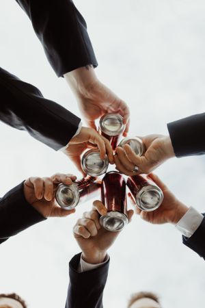 Groomsmen in dark suits toasting with canned drinks photographed from below in a circular huddle.