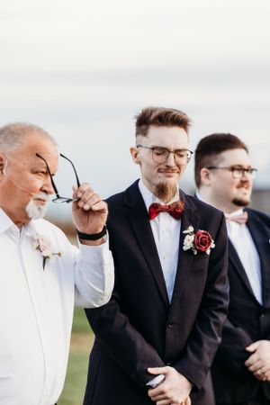Emotional older man removing glasses beside smiling groom in navy suit with red rose boutonniere.