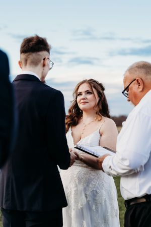 Bride in lace gown gazing at groom during outdoor wedding ceremony officiated at sunset.