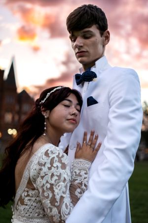 Elegant couple in white wedding attire embracing at dusk under a dramatic colorful sunset sky outdoors.