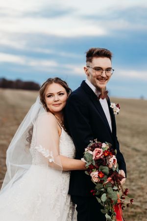 Smiling groom in navy suit with bow tie and bride holding cascading red and pink bouquet in a field.